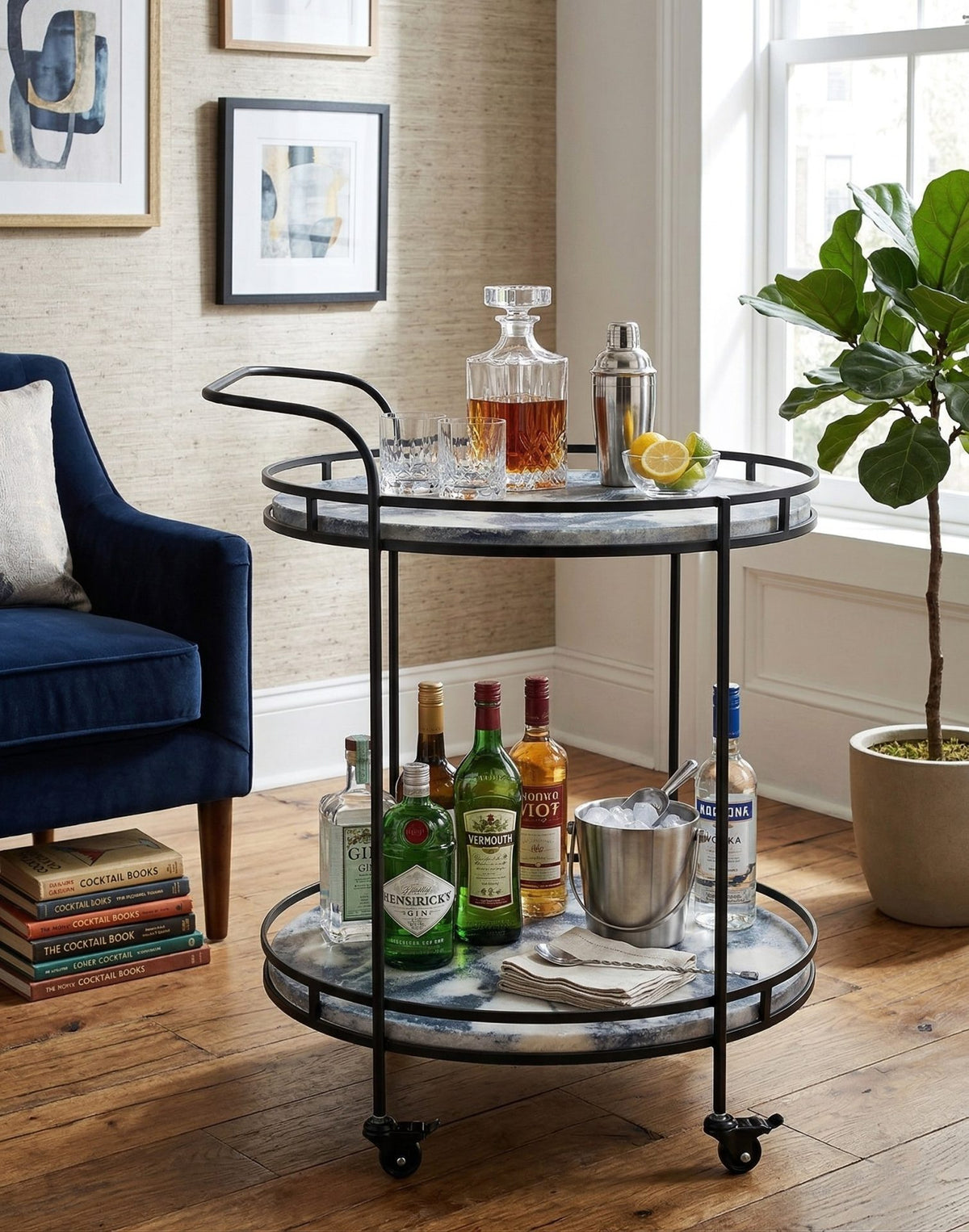 A two-tier bar cart with a black metal frame and a white-and-gray marbled tray, styled in a bright living room with bottles, glasses, and bar tools on top.